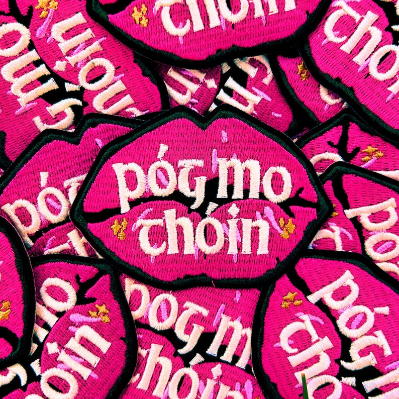 May include: Pink fabric patches with the text "p&oacute;g mo choin" in white lettering. The patches are shaped like lips and have a black outline.