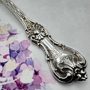 May include: Silverware handle with ornate details and an engraved monogram. The handle is silver and has a floral design.