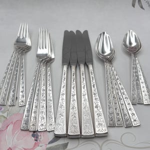 May include: An assortment of silver-coloured cutlery, including forks, knives, and spoons. The handles are decorated with an engraved floral pattern. The arrangement suggests a formal dining setting.