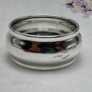 Op de afbeelding: Een zilveren servetring met een decoratief bloemmotief. De ring is rond en heeft een gladde, gepolijste afwerking.