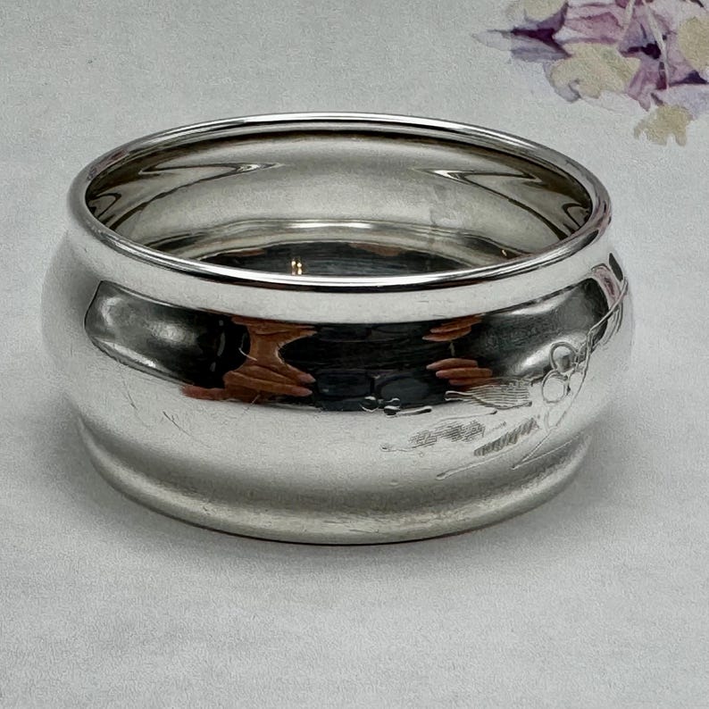 Op de afbeelding: Een zilveren servetring met een decoratief bloemmotief. De ring is rond en heeft een gladde, gepolijste afwerking.