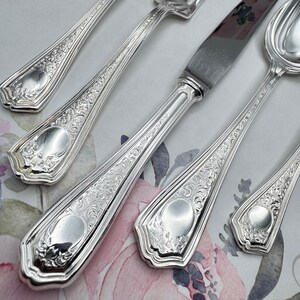 Peut inclure: Gros plan sur un ensemble de couverts en argent ornés, comprenant des fourchettes, des couteaux et des cuillères. L'argenterie présente des motifs floraux et de volutes détaillés. Le métal poli reflète la lumière, soulignant les motifs complexes.