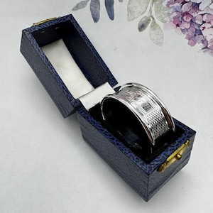 May include: A silver napkin ring with a woven design, in a blue velvet box. The box is open and the ring is inside.