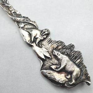 Peut inclure: Un manche de cuillère souvenir en métal argenté avec un motif en relief. Le manche présente le mot ALASKA en lettres en relief, un morse et un ours polaire debout sur un paysage glacé avec des détails texturés sur tout le métal.