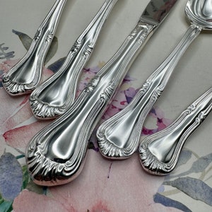 May include: A close-up shot of a collection of silver-coloured cutlery. The set includes spoons, forks, and a knife, each piece featuring an elaborate, flowing design along the handles. The reflective surfaces create highlights and shadows, emphasising the intricate patterns.