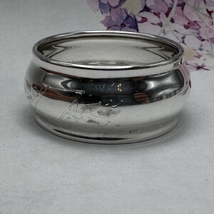 Op de afbeelding: Een zilveren servetring met een gegraveerd bloemenmotief. De ring is gemarkeerd met "STE" aan de onderkant.