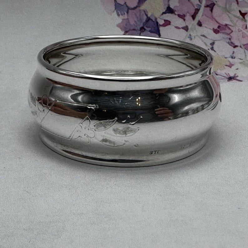 Op de afbeelding: Een zilveren servetring met een gegraveerd bloemenmotief. De ring is gemarkeerd met "STE" aan de onderkant.