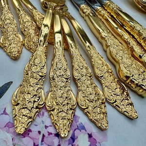 May include: Close-up of ornate gold-coloured silverware, including forks and knives. The handles feature detailed floral patterns. The silverware is arranged on a surface with a soft, blurred floral design in shades of purple and white.