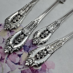 May include: Close-up of ornate silver cutlery. The cutlery features a floral design with roses in the centre of the handles. The handles are framed with a decorative border. The cutlery is arranged on a floral patterned surface.