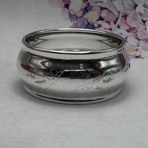 Op de afbeelding: Een zilveren servetring met een gegraveerd ontwerp in de vorm van een diamant met bloemmotieven.