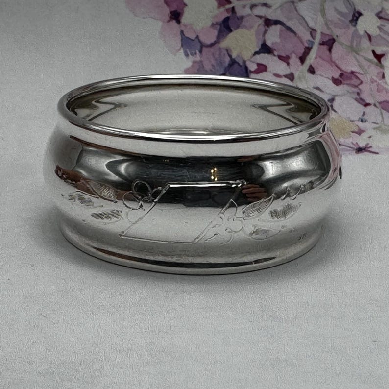 Op de afbeelding: Een zilveren servetring met een gegraveerd ontwerp in de vorm van een diamant met bloemmotieven.