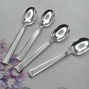 May include: Four silver spoons with oval bowls and ridged handles are arranged diagonally. The spoons have a shiny, reflective surface, and the handles have a rectangular base. The spoons are likely for serving or eating.