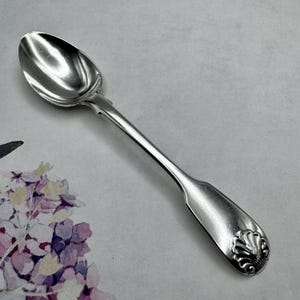 May include: A silver teaspoon with an ornate handle and a rounded bowl. The handle features a decorative shell design.