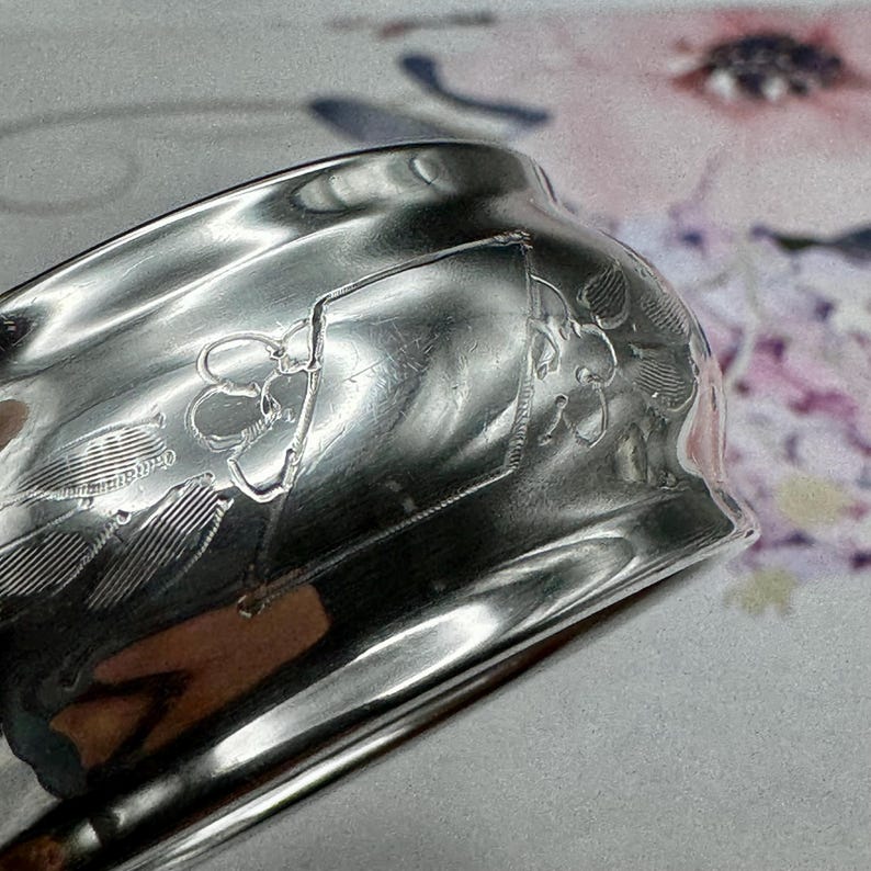 Op de afbeelding: Een zilveren servetring met een ingewikkeld bloemmotief. De ring is gepolijst en glanzend en reflecteert het licht.