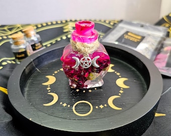May include: A small pink glass bottle with a cork stopper and a silver charm. The bottle is filled with dried rose petals and a clear liquid. The bottle is sitting on a black tray with a gold crescent moon design.