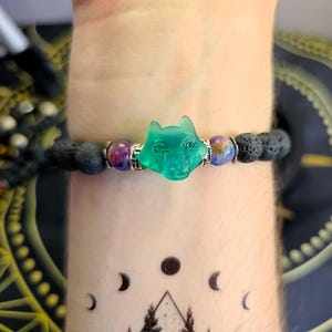 Puede incluir: Un brazalete de roca de lava negra con un dije de lobo verde y cuentas de piedras preciosas moradas y azules.