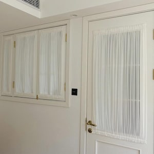 以下が含まれることがあります： 窓とドアに掛けられた白いシアーカーテン。カーテンは軽くて流れるような生地で作られています。窓とドアのフレームは白で、金具は金色の色合いです。部屋は明るく照らされています。