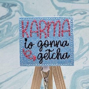 May include: A blue glitter badge holder with the text "Karma is gonna getcha" in pink and black embroidery. The badge holder is on a small wooden easel.