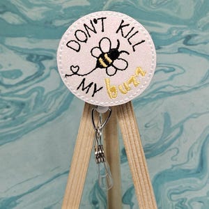 May include: White round badge with a black and yellow bee and the text "Don't Kill My Buzz" in black and gold lettering. The badge is attached to a silver retractable badge reel.