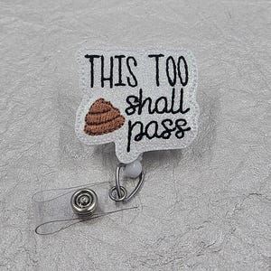 May include: A white glittery badge reel featuring an embroidered brown poop emoji and the black stitched text "THIS TOO shall pass". The badge reel includes a clear plastic strap with a metal snap button and a silver metal swivel clip attachment.