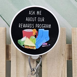 May include: A black and white round badge with the text "Ask me about our rewards program" and a cartoon image of a coffee cup, socks, a sandwich, and other items. The badge is attached to a clear plastic retractable badge holder.