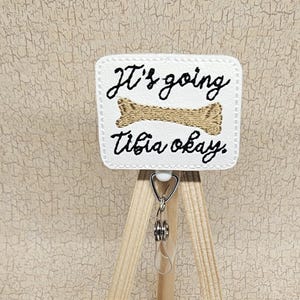 May include: White square sign with black text that reads "It's going tibia okay."  A gold bone is pictured above the text. The sign is on a small wooden easel.