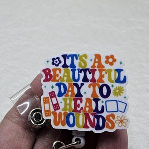 May include: A colorful badge reel with the text "IT'S A BEAUTIFUL DAY TO HEAL WOUNDS" in a retro font. The design includes images of a bandage and a medical kit. The reel has a clear plastic strap and a metal clip.