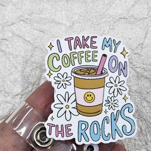 May include: A badge reel with a colorful design featuring a coffee cup with the text "I take my coffee on the rocks." The cup has a smiley face and is surrounded by daisies and stars.