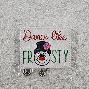 May include: A white badge reel with the text "Dance like Frosty" in red and green. The design features a snowman with a black hat and a pink flower. The badge reel has a clear plastic strap and a metal clip.