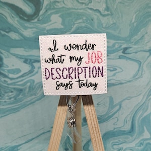 May include: White square badge with the text "I wonder what my JOB DESCRIPTION says today" in black, pink, and purple embroidery. The badge is attached to a silver retractable badge holder.