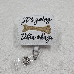 May include: A white rectangular badge reel featuring black embroidered text that reads It's going Tibia okay. Centered between the lines of text is a gold embroidered bone graphic. The badge reel includes a clear plastic strap with a metal snap button.