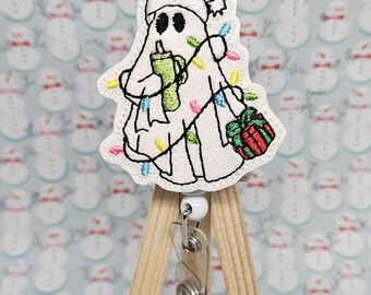 May include: A white ghost wearing a red Santa hat and holding a green cup is decorated with colorful lights and a red gift. The ghost is attached to a wooden retractable badge holder.