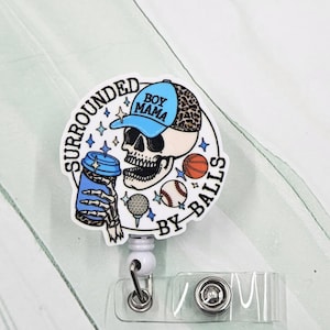 May include: A white badge reel with a skull wearing a blue and leopard print hat, holding a blue coffee cup with a skeleton hand. The text reads "SURROUNDED BY BALLS" and "BOY MAMA". Includes a basketball, golf ball, and baseball.