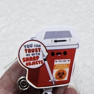 May include: A red and white badge reel featuring a cartoon sharps container with a syringe. The badge reel has the text "YOU CAN TRUST ME WITH SHARP OBJECTS" and "WARNING BIOHAZARD".