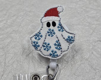 May include: A white ghost badge reel featuring embroidered blue snowflakes across its body and a red and white Santa hat on its head. The ghost has two black oval eyes and is attached to a clear plastic strap with a metal snap button fastener.