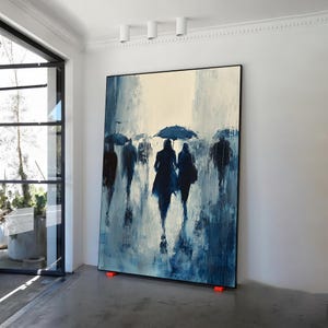 May include: A large abstract painting of people walking in the rain. The painting is done in shades of blue and white, with the figures silhouetted against a light background. The painting is framed in black and is hanging on a white wall.