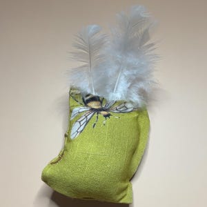 Puede incluir: Una pequeña bolsa de tela verde lima con una ilustración de abeja y plumas blancas. La bolsa tiene un aspecto texturizado y un ribete dorado. El diseño de la abeja presenta una abeja amarilla y negra con alas detalladas.