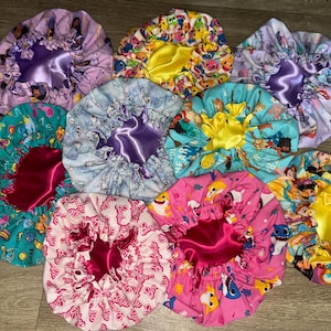 May include: A selection of patterned satin bonnets. The bonnets feature cartoon characters and bow designs. The bonnets have a contrasting satin lining in pink, purple, and yellow. These bonnets are designed to protect hair and maintain hairstyles.