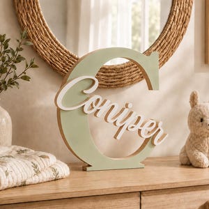 May include: A large, sage green wooden letter C featuring the white cursive name Coniper layered across the front. The decorative piece has a flat base for standing and is finished with a smooth, matte texture, showcasing a clean, modern design.