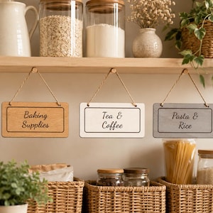 Può includere: Tre targhe rettangolari in legno da appendere con angoli arrotondati e cordini. La targa a sinistra è marrone chiaro con il testo Baking Supplies, quella centrale è bianca con Tea & Coffee e quella a destra è grigia con Pasta & Rice.