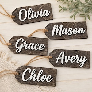 Könnte beinhalten: Fünf rechteckige, dunkelbraune Holzanhänger mit abgerundeten Ecken, jeweils mit einem ausgeschnittenen Namen in weißer Schreibschrift. Die Namen lauten Olivia, Mason, Grace, Avery und Chloe. Jeder Anhänger hat eine Schlaufe aus Jutekordel.