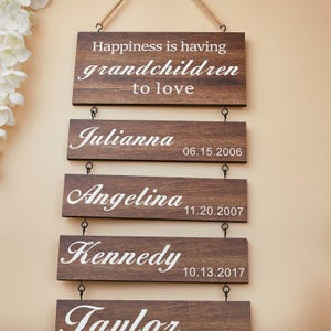 May include: A wooden family sign with the text "Happiness is having grandchildren to love." Below are the names Julianna, Angelina, and Kennedy, with their birthdates. The sign is made of dark brown wood and has a rope for hanging.