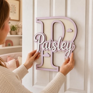 Pode incluir: Uma letra P grande de madeira em lilás claro e creme com o nome Paisley escrito em letra cursiva branca no centro. O sinal apresenta pequenos recortes de nuvens e está montado numa porta branca, com um design em camadas.