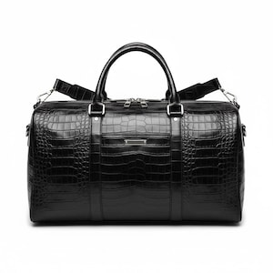 May include: Black duffel bag with a crocodile skin effect. The bag features a top handle and a detachable shoulder strap. Silver-toned hardware, including zippers and strap attachments, adds detail. A small, rectangular metal plate is centered on the front.