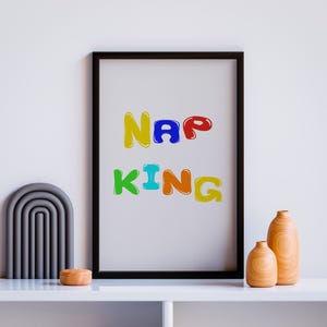 May include: A framed print with the text "NAP KING" in colorful, bubble-like letters. The letters are in shades of red, orange, yellow, green, blue, and purple. The print is hanging on a white wall above a shelf with two wooden vases and a gray arch-shaped object.