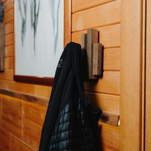 May include: A wooden wall-mounted coat rack with three vertical wooden pegs. A black quilted jacket hangs from the rack. The wall is made of horizontal wooden planks, and artwork is visible in the background.