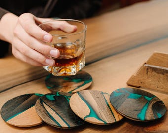 May include: Set of five wooden coasters with a unique design. Each coaster features a combination of natural wood and vibrant teal resin. The coasters are oval-shaped and sit on a wooden surface. A glass of amber liquid is held above the coasters.
