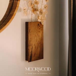 Op de afbeelding: Een houten wandvaas met een droogbloemenarrangement. De vaas is gemaakt van gerecycled moeras eikenhout en heeft een natuurlijke houtkleur. De tekst "MOORWOOD - BOG OAK IS ETERNAL -" is onder de vaas gedrukt.