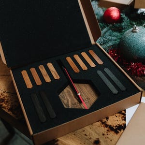 May include: A wooden clock kit presented in a cardboard box with a black foam insert. The kit includes a hexagonal clock face, wooden clock hands, and various wooden pieces in different shades. The clock hands are red.