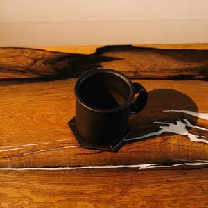 May include: A black ceramic mug with a handle sits on a wooden table with white streaks. The mug is filled with a dark liquid.